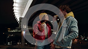 Positive couple eating snacks food at night. Man feeding woman under bridge.