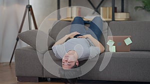 Positive creative Asian young man thinking lying upside down on couch with laptop. Wide shot portrait of inspired