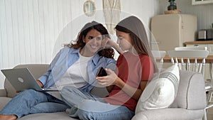 Positive curious mother with laptop and shy teenage girl with mobile phone sits on sofa in home