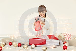 Positive cute female kid wearing white sweater holding soft toy sitting on bed with Christmas decoration and garland, looking at