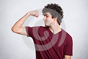 Positive dark haired teenager, dressed casually, shows biceps with satisfied facial expressions, demonstrates how cool he is.