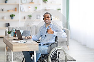 Positive disabled black man in headset using laptop computer for online work, showing thumb up gesture at home