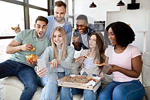 Positive diverse friends having home party, sitting on couch with pizza, chips and smartphone, having fun together