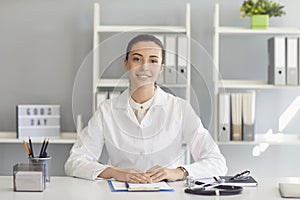 Positive doctor woman looking at the camera smiling while sitting at a table in a clinic.