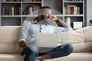 Positive dreamy African American man relaxing on couch with laptop