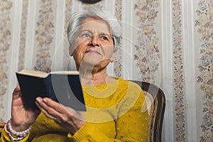 Positive elderly retired woman sitting on a chair, slowly reading a book and daydreaming. Peaceful retirement