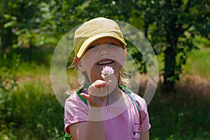 Positive emotional young lady smiles, looks on wild flower that holds in hand. Cheerful pretty child girl in yellow cap