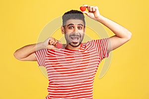 Positive excited bearded man in striped t-shirt fooling around having fun with red toy hearts, showing his fondness and devotion, photo