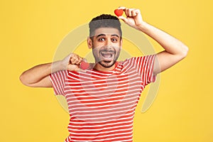 Positive excited bearded man in striped t-shirt fooling around having fun with red toy hearts, showing his fondness and devotion,