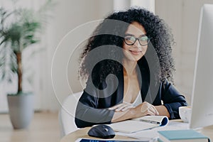Positive female director with Afro curly hair, rewrites information in notepad from computer, wears optical glasses and formal