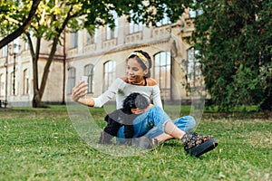 Positive female dog owner sitting on grass with pet and taking selfie on smartphone camera with smile on face. Photo with a dog on