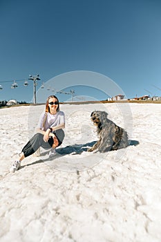 Positive female hiker in casual clothes sitting on snow in mountains and posing at camera with smile on face near dog