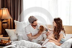 Positive female and male have fun together with their dog in bed, enjoy calm domestic atmosphere and togetherness