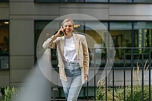 Positive female office worker talking phone while standing on modern office terrace and looks camera