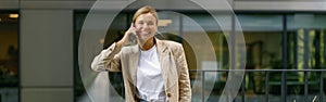 Positive female office worker talking phone while standing on modern office terrace and looks camera
