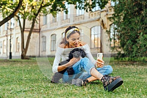 Positive female owner sitting with a little black dog on the grass and taking a selfie with a smile on her face. Beautiful lady