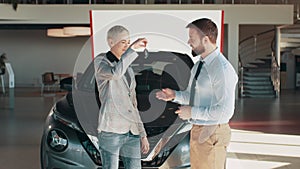 Positive female in smiling for camera and demonstrating keys while standing near new vehicle in showroom. Car salesman