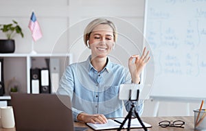 Positive female teacher giving online English lesson on smartphone, showing OK gesture at home photo