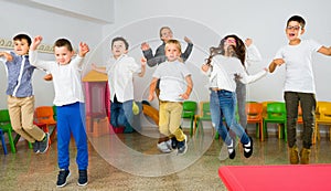 Positive female teacher and group of schoolkids jumping in classroom
