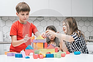 Positive, focused children of boy and two girls playing with colorful blocks and building castle from geometric figures