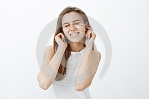 Positive funny european woman in casual tank-top, closing ears with index fingers and smiling broadly with closed eyes