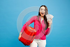 Positive girl with baggage and money are going to travel. Woman in pink sweater and jeans standing on blue background. Vacation