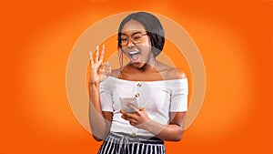Positive Girl Holding Phone Gesturing Okay And Winking, Orange Background
