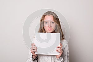 Positive girl holds a blank poster for text. Schoolgirl with a smile holds a white sheet for text