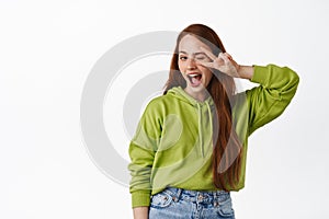 Positive girl with long natural red hair, show peace v-sign gesture near winking eye, smiling happy at camera, posing in