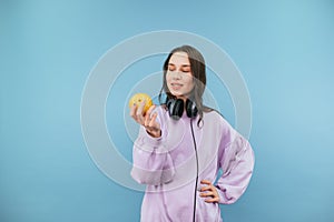 Positive girl in sweatshirt and headphones around her neck isolated on blue background with apple in hands, looks at fruit and