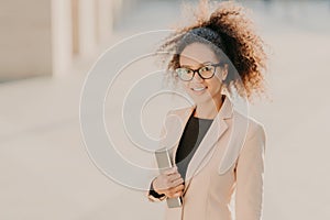 Positive gorgeous Afro American businesswoman strolls outdoor with touchpad, wears formal clothes, optical spectacles, smiles at