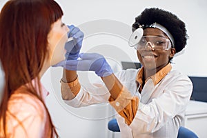 Positive grinning young African American lady doctor otorhinolaryngologist, wearing uniform, frontal reflector and