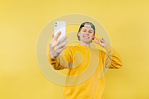 Positive guy in a cap and yellow sweatshirt takes a selfie on camera and smartphone and shows a gesture of peace and winks