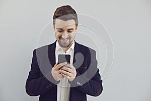 Positive guy in a jacket is typing text in a mobile smiling with on a gray background.