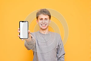 Positive guy shows a modern smartphone with a white screen, looks at the camera and smiles on a yellow background. Smartphone with