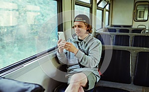 A positive guy with a smartphone in his hands sits in a train by the window while traveling and uses the phone with a smile on his