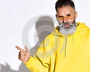 Positive handsome bearded middle aged man in yellow hoodie and glasses standing and pointing aside with finger