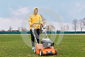 Positive handsome guy in yellow hoodie with gasoline mower in his hands, man mows grass on lawn, improvement of territory, male