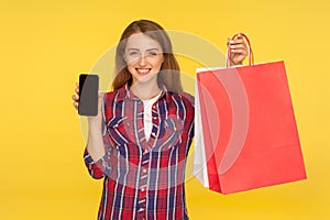 Positive happy good-natured ginger girl in casual style checkered shirt smiling, holding packages and mobile phone