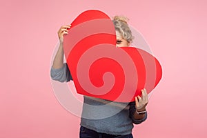 Positive happy woman with curly hair hiding face behind big red heart and looking at camera with curious prying eyes