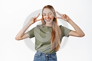 Positive and happy young blond girl, winking and smiling white teeth, showing peace v-sign gesture near eyes, standing