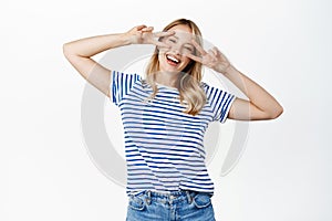 Positive and happy young woman, 25 years old, laughing and smiling, showing v-sign, peace gesture near face, standing