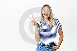 Positive and happy young woman, 25 years old, laughing and smiling, showing v-sign, peace gesture near face, standing