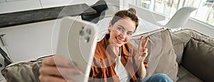 Positive and happy young woman taking selfie on smartphone, sitting on couch, showing peace, v-sign gesture