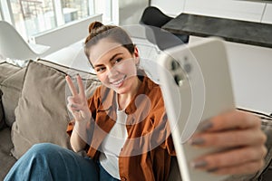 Positive and happy young woman taking selfie on smartphone, sitting on couch, showing peace, v-sign gesture