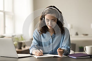 Positive hardworking online student girl in headphones writing summary notes