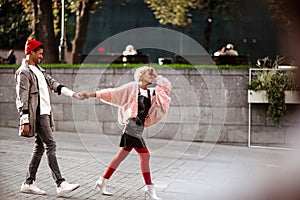 Positive hipster guy and his mulatto girlfriend hold hands and walk together in street, being in good mood, enjoy their