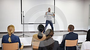 Positive Hispanic woman standing with microphone on stage in conference room, speaking to businesspeople at seminar