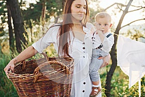 Positive housewife with hangs white clothes to dry. Young mother with her little son is outdoors in the forest. Beautiful sunshine