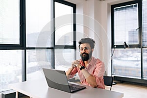 Positive Indian businessman in casual clothes sitting at desk with laptop smiling looking at camera in light coworking